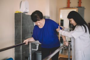 Caring asain chinese female physical therapist helps senior woman stroke victim in rehab center walking with the help of parallel bars